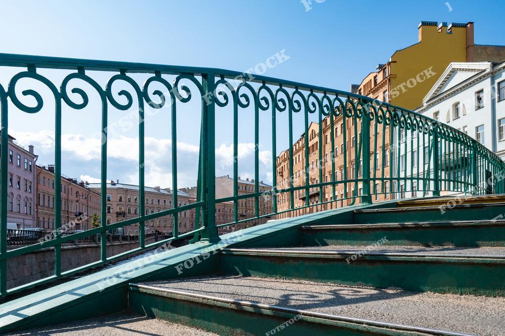 На фотографии изображен пешеходный мост с изящными коваными перилами, переброшенный через один из каналов Санкт-Петербурга. На заднем плане видны фасады исторических домов, отражающие классическую архитектуру города. Яркий солнечный день подчеркивает богатую палитру красок городской среды. Этот вид характерен для районов набережных, придающих Петербургу его неповторимый облик.